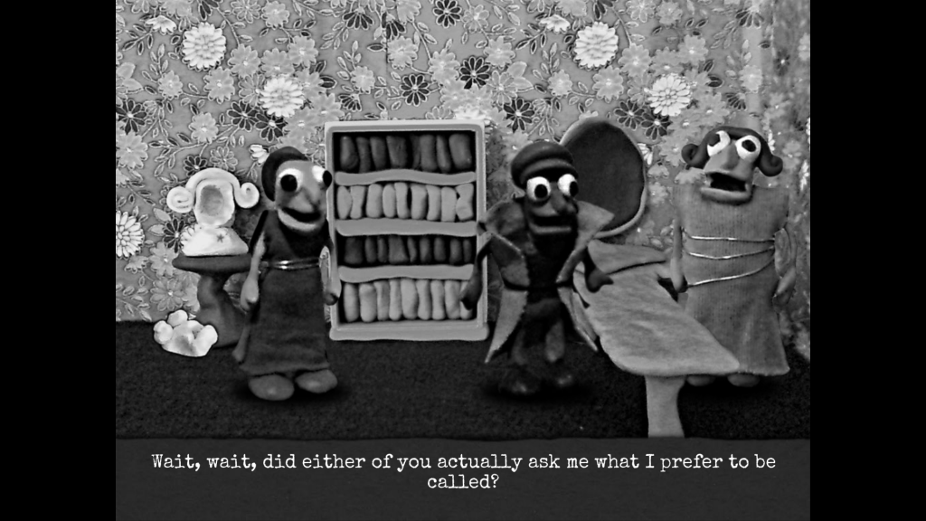 Three clay figures (Dominique and two others) in a floral wallpapered office with desk, full bookshelf and broken bust statue, one is behind the desk, Dominique and another in a dress in front. Dominique gestures in frustration, text reads, 'Wait, wait, did either of you actually ask what I prefer to be called?'