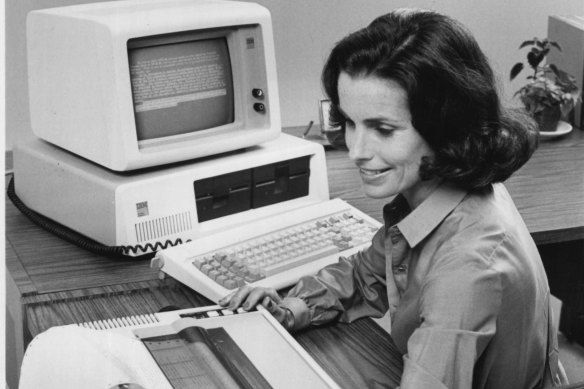 several decades old picture of woman sat at computer desktop smiling 