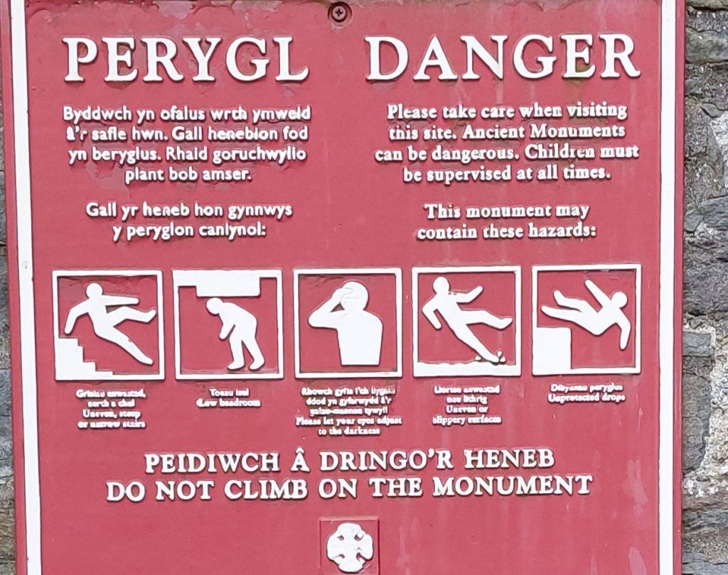 a sign in welsh and english explaining the dangers of navigating a historical site, with pictures of a figure in different types of peril appropriate to context