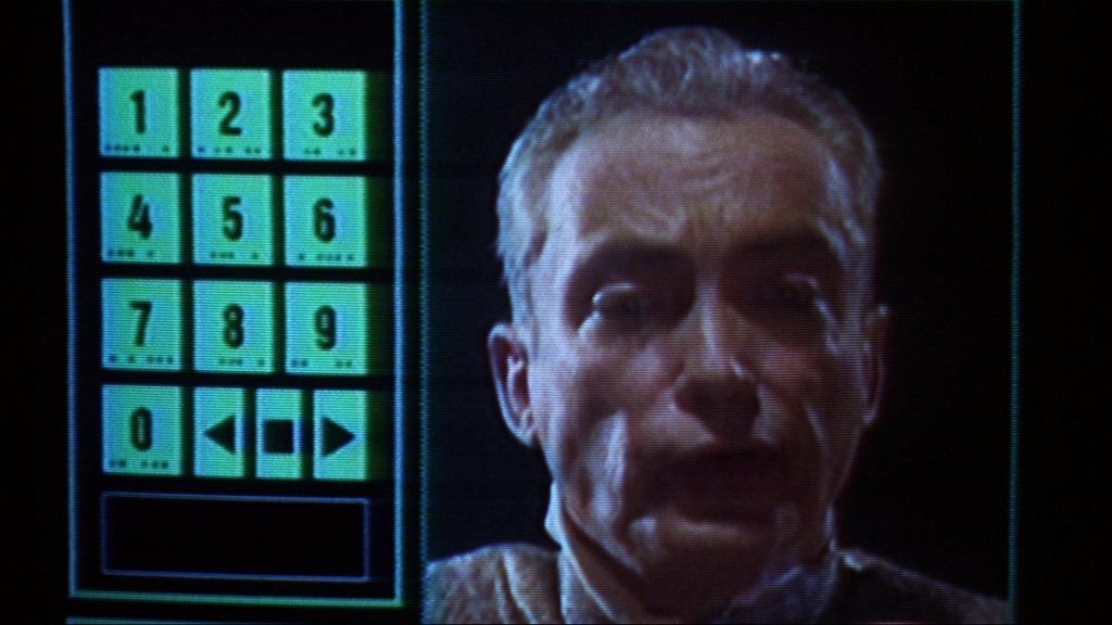 udo kier looking on a videocall with a number pad besides his head 
